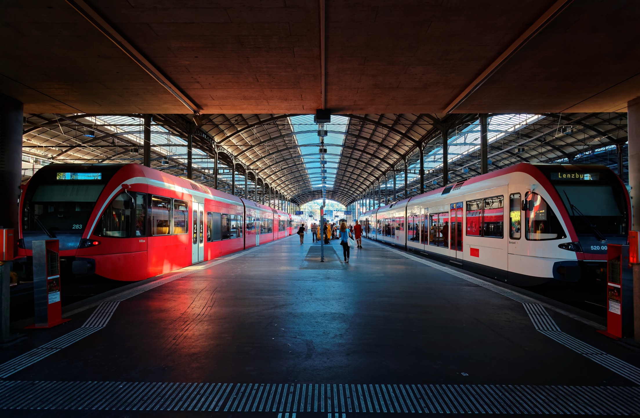 An island train station platform with one train on either side and passengers moving about.