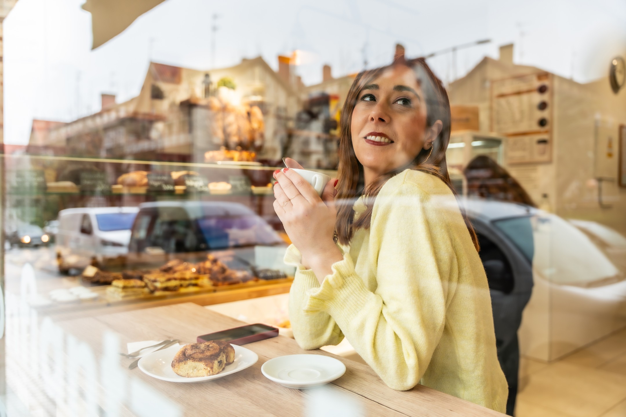 A woman sits in front of the window of a cafe, looking out towards the street.